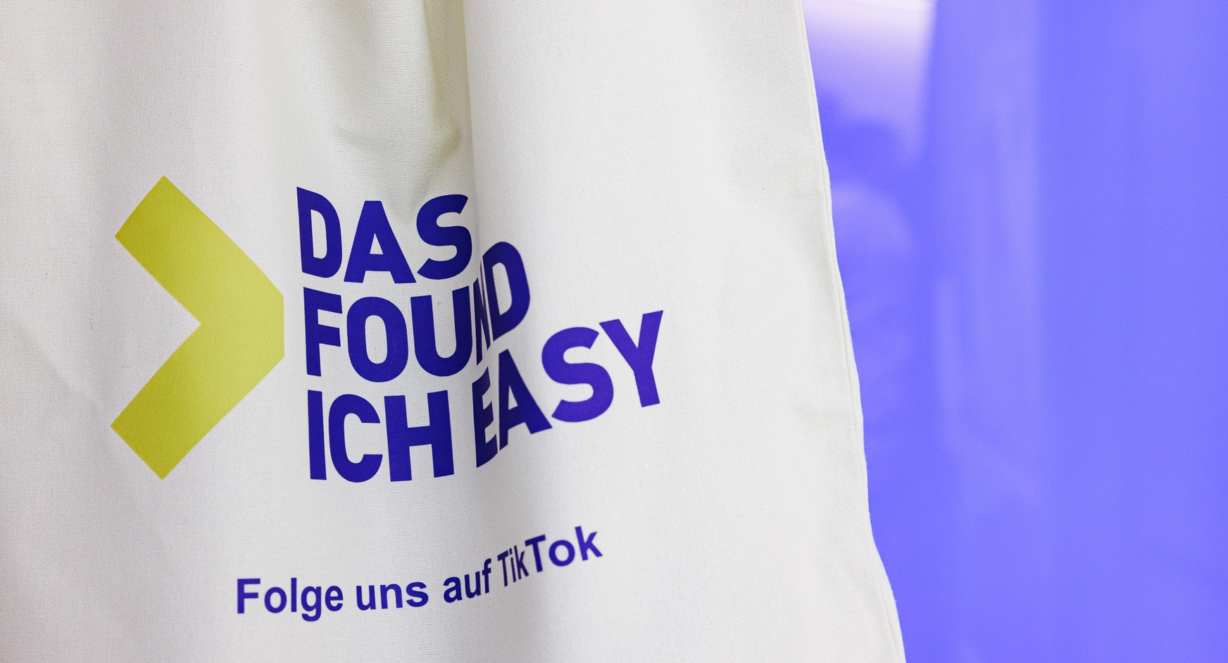 Naufnahme einer weißen Jutetasche vor violett-floriszierendem Hintergrund. Auf der Tasche steht der Schriftzug Das found ich easy Folge uns auf TikTok