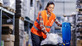 Person mit geschlossenen dunklen Haaren und kariertem Hemd sowie roter Warnweste entsorgt Müll in einer blauen Tonne in einer Lagerhalle