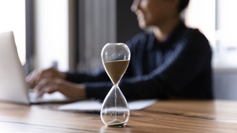 Eine Sanduhr steht auf einem Tisch. Die Sandkörner fließen durch die Öffnung. Im Hintergrund sitzt eine Person am Tisch. Sie blickt in einen aufgeklappten Laptop und hat ihre Hände auf der Tastatur liegen.