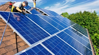 Person bei der Montage von Solarpanelen auf Hausdach unter blauem Himmel mit Wolken, im Hintergrund grün belaubter Baum