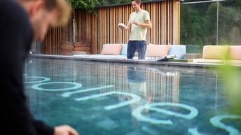 Person mit kurzen dunklen Haaren, grünem T-Shirt und Jeans steht bei einem Beckenrand eines beheizten Outdoor-Pools und hält ein Gerät in der Hand, am vorderen linken Bildrand steht eine weitere Person in Unschärfe am Beckenrand