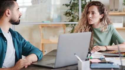 Personen sitzen an einem Schreibtisch und unterhalten sich in einem Büro, ein Laptop und weitere Unterlagen liegen am Tisch