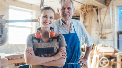 Zwei lächelnde unterschiedlicher Generationen in Arbeitsgewändern stehen in Tischlerwerkstatt, eine Person hat Schutzbrille am Kopf, Ohrenschützer um den Hals und hält die Arme verschränkt