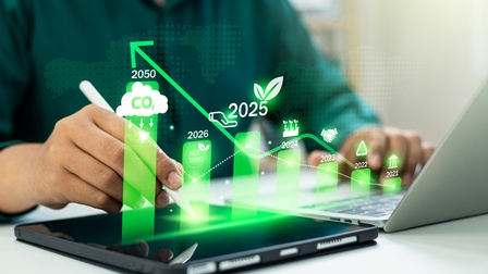 Nahaufnahme einer Person, die auf einem Laptop tippt, sie hat einen Stift in der Hand, daneben liegt ein Tablet. Man sieht ein grünes Balkendiagramm mit den Zahlen 2025 und 2025 und Symbolen: Wolke mit Co2, Blätter, Kreislauf, Parlament, Hände.