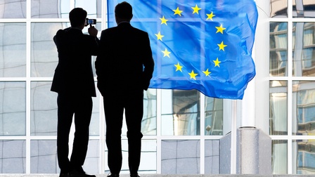 Personen in Businesskleidung stehen vor einem Gebäude mit EU-Flagge im Schatten, eine Person fotografiert dabei die Flagge