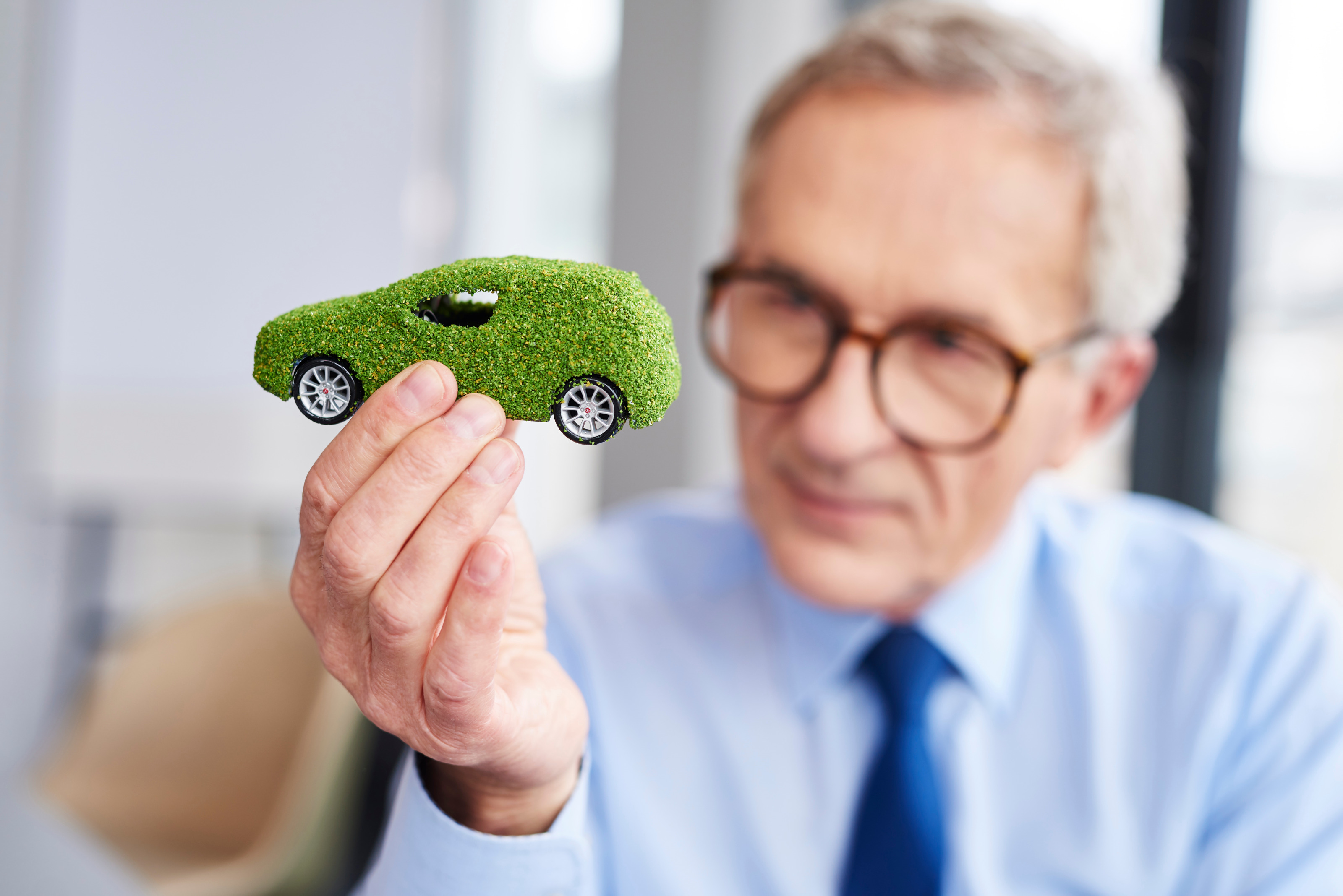 Eine Person mit Brille und blauem Hemd hält ein kleines Miniaturauto in die Kamera. Das Auto ist mit grünen, unebenen Stoff überzogen.