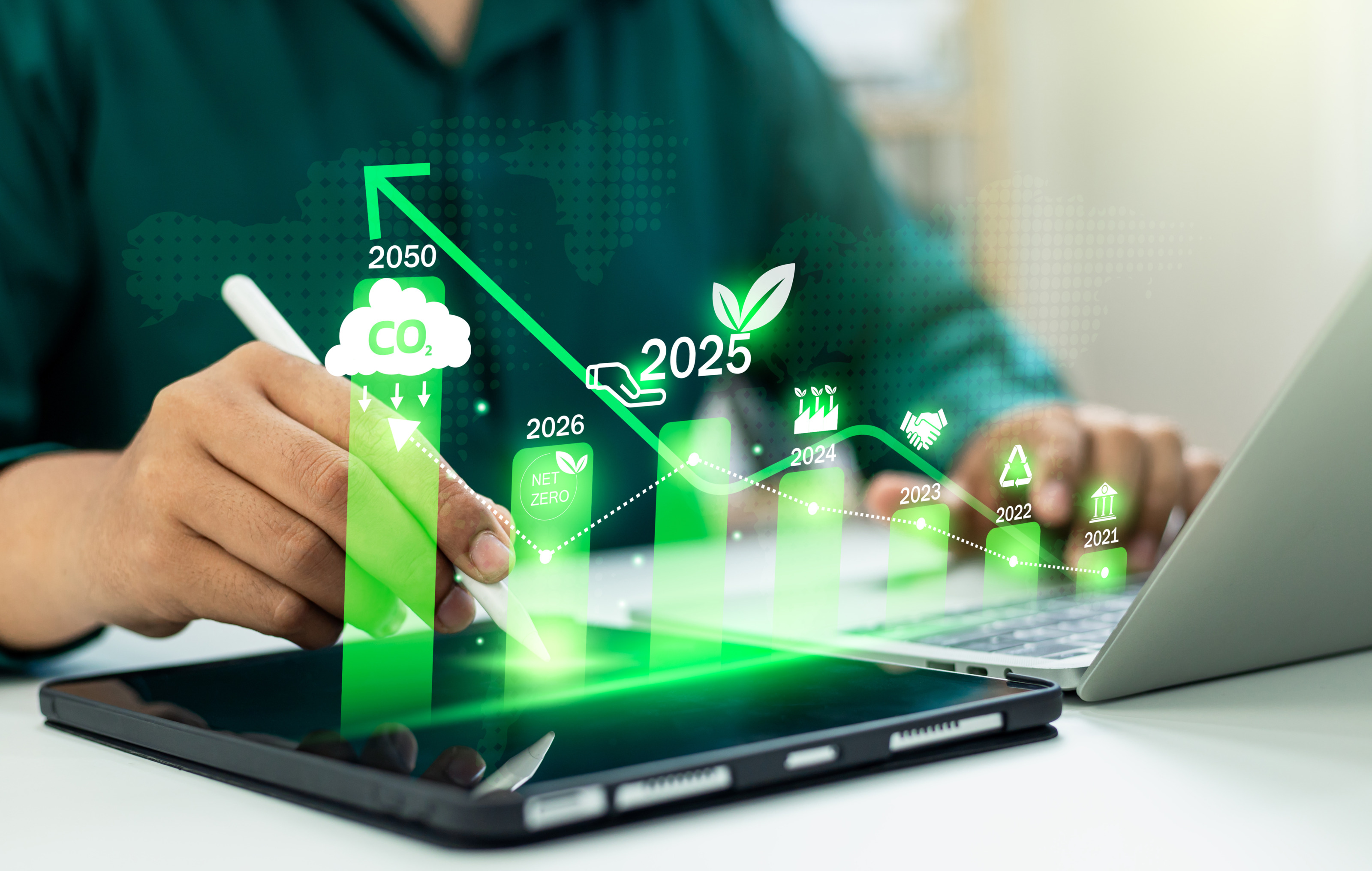 Nahaufnahme einer Person, die auf einem Laptop tippt, sie hat einen Stift in der Hand, daneben liegt ein Tablet. Man sieht ein grünes Balkendiagramm mit den Zahlen 2025 und 2025 und Symbolen: Wolke mit Co2, Blätter, Kreislauf, Parlament, Hände.